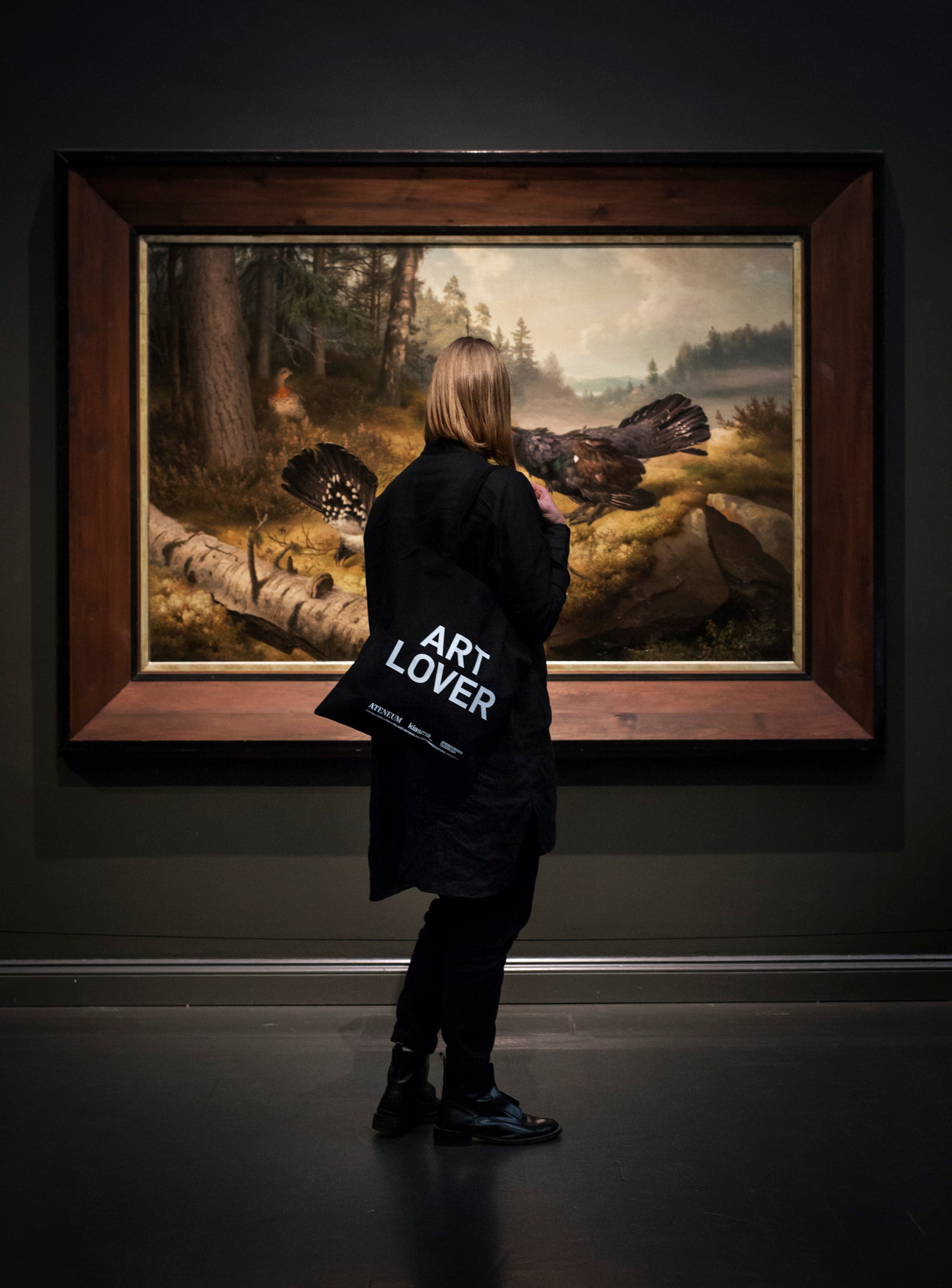 A photograph of a person looking at a painting depicting capercaillies, with a canvas bag on their shoulder that reads "Art lover."
