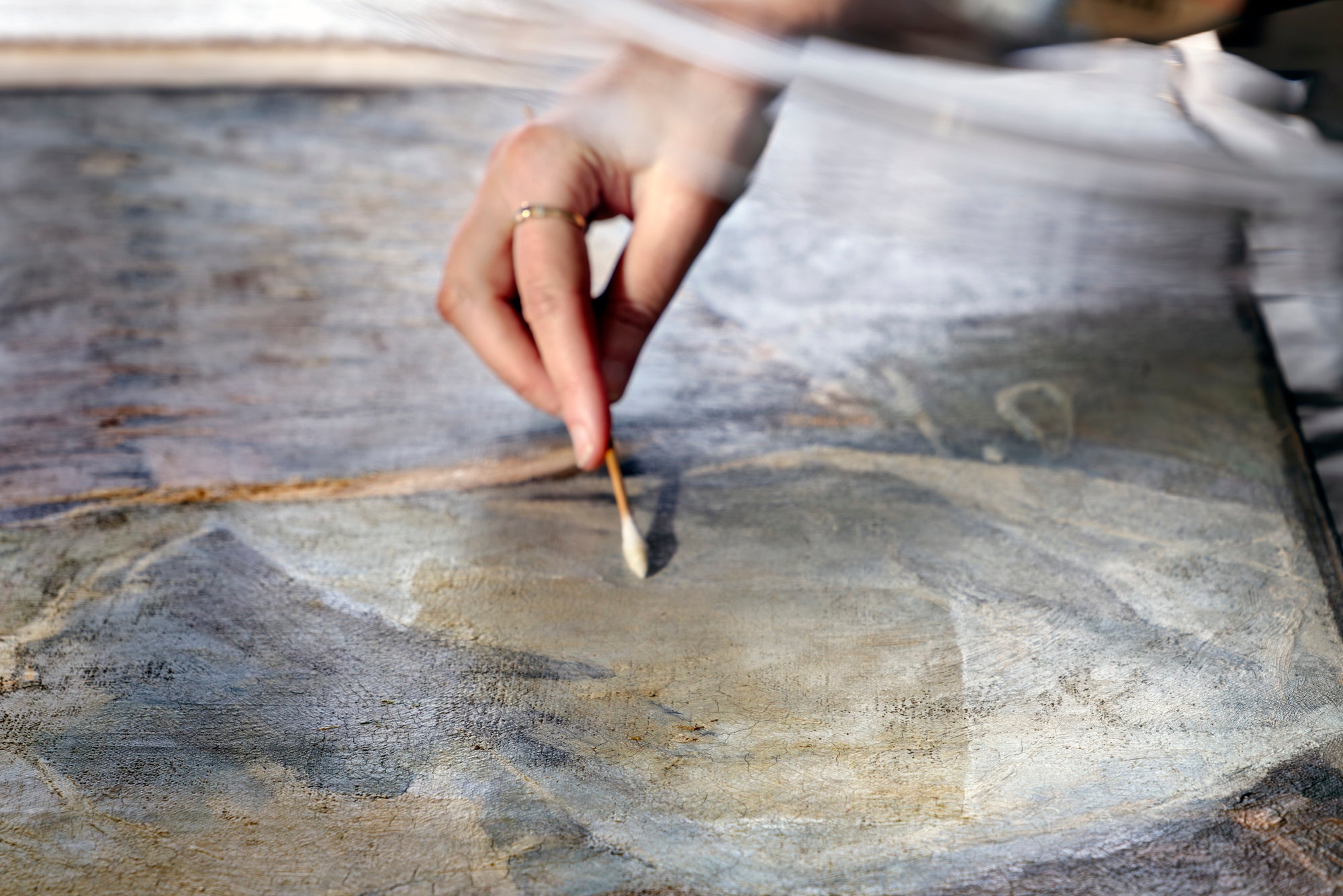 The photograph shows a hand gently stroking the surface of an oil painting with a cotton swab.