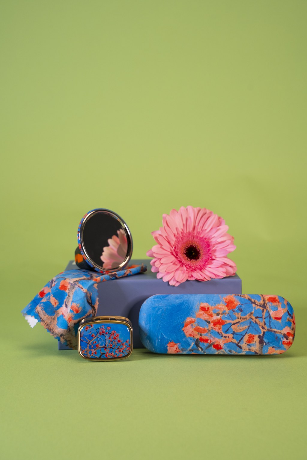 The photograph shows products made from the same artwork depicting a flowering tree: a pillbox, a glasses case, and a small mirror.