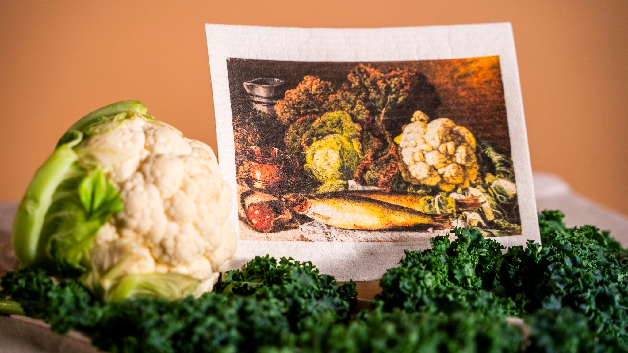 A photograph of a dishcloth featuring a still life painting of vegetables. Alongside the dishcloth, the photo shows the same vegetables as in the painting: cauliflower and kale.
